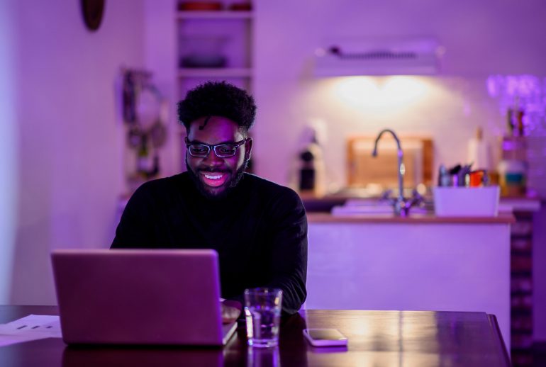 A cheerful african american freelancer is sitting at home at night and working on a project with cutting-edge technologies. An e-commerce worker is typing on a laptop and finishing his work at night.